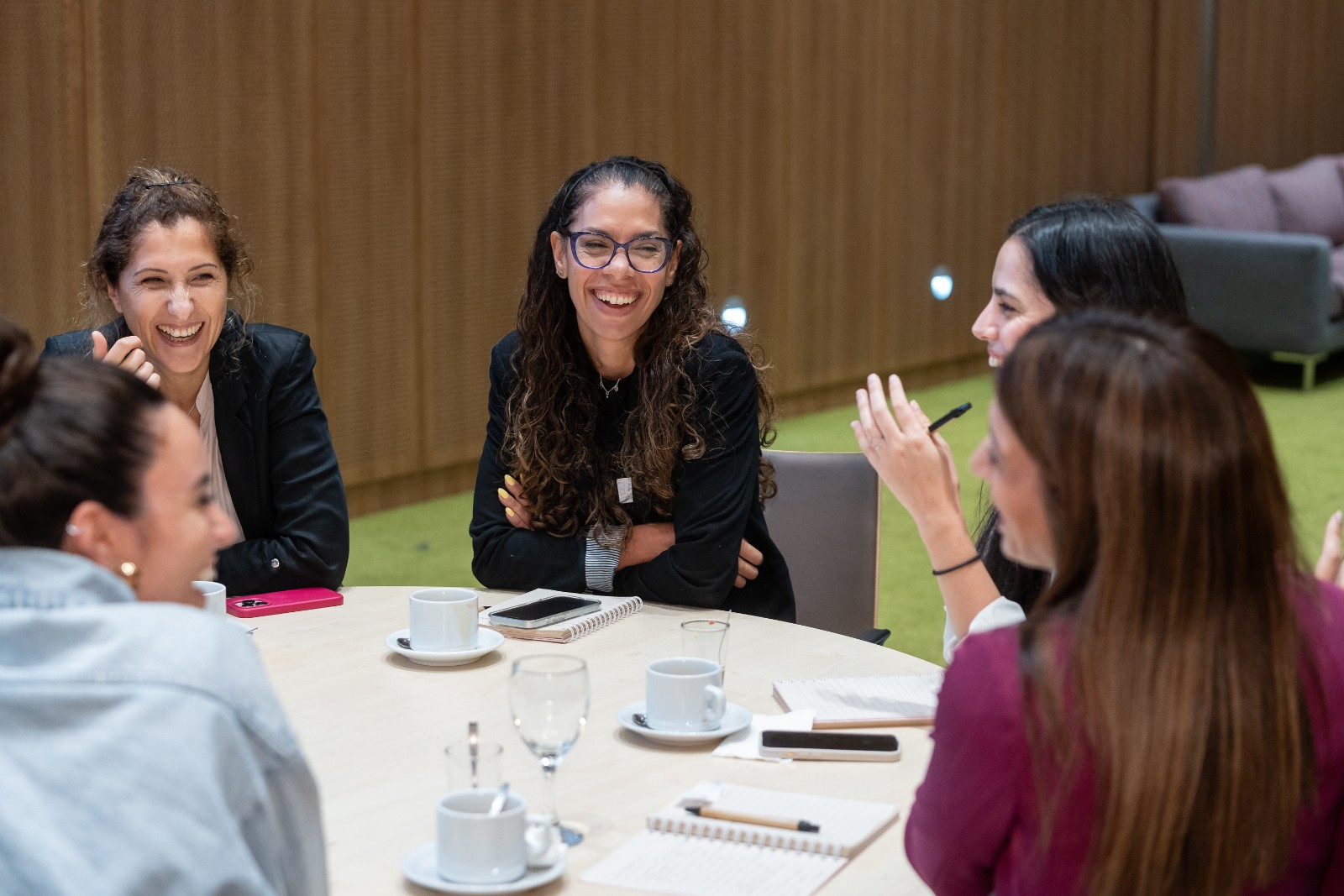Un grupo de 5 mujeres hace networking y ríe en una mesa de trabajo.