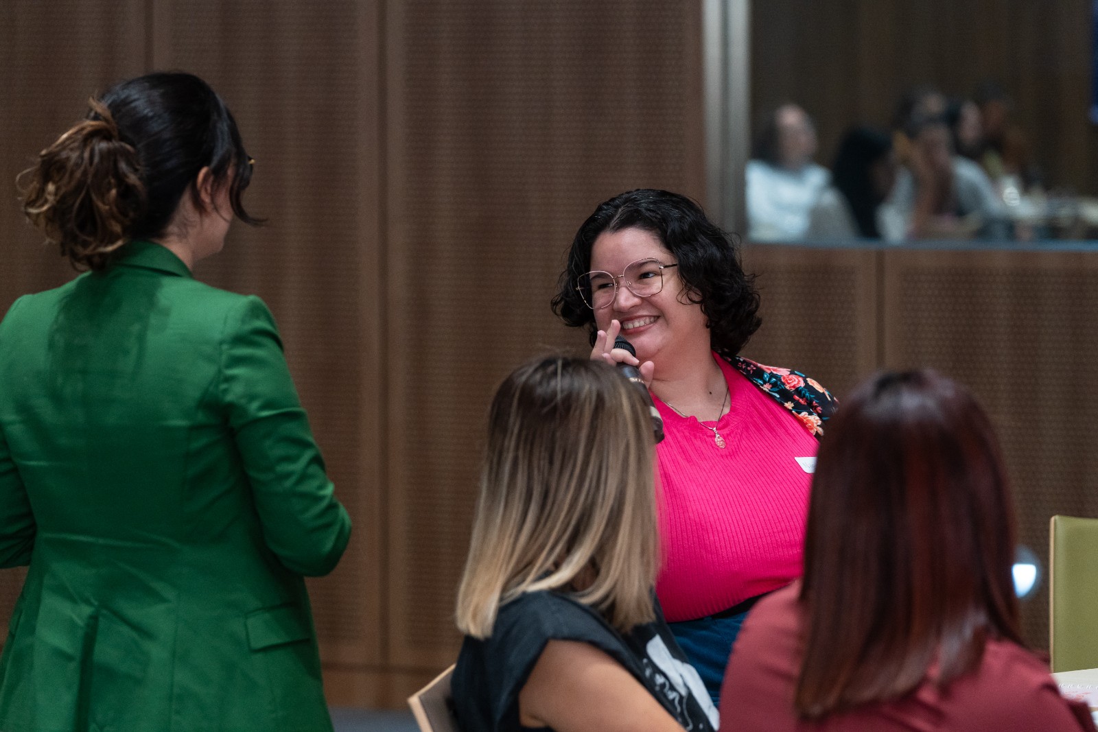 Melanie Tobal (speaker con blazer verde), se acerca a una participante en una mesa, quien sostiene un micrófono y sonríe.